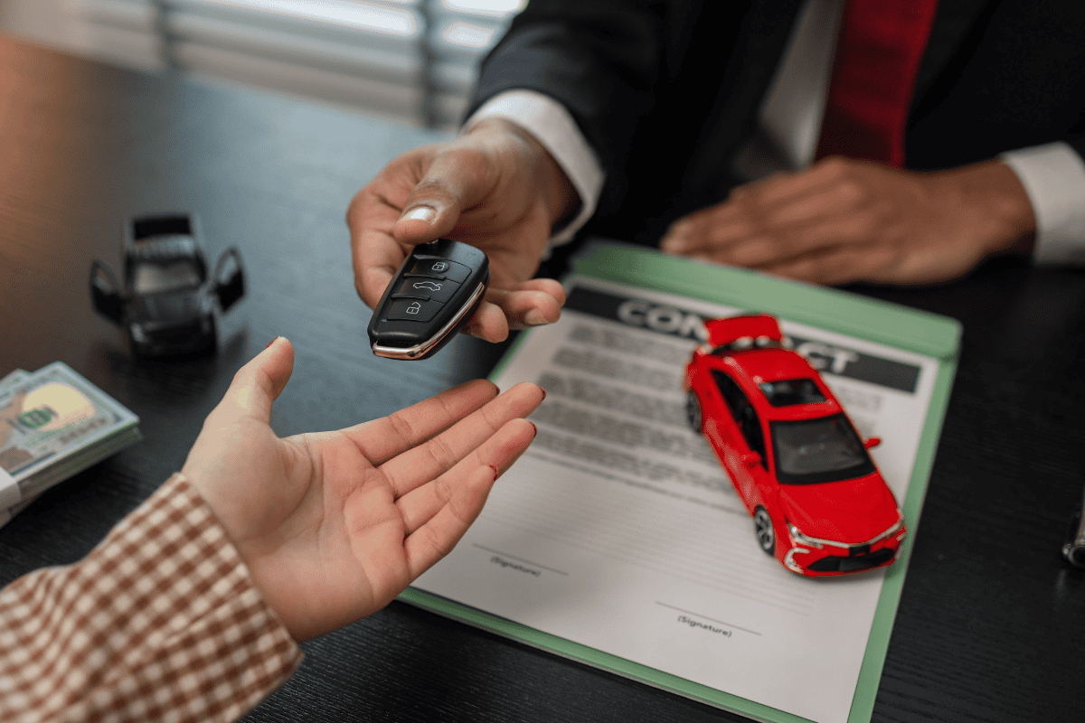 Dealer hands over car key to customer while a red car model sits on a contract on the desk