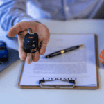 Close-up of a car key fob and keys held over a contract on a clipboard, with a toy car and laptop nearby.
