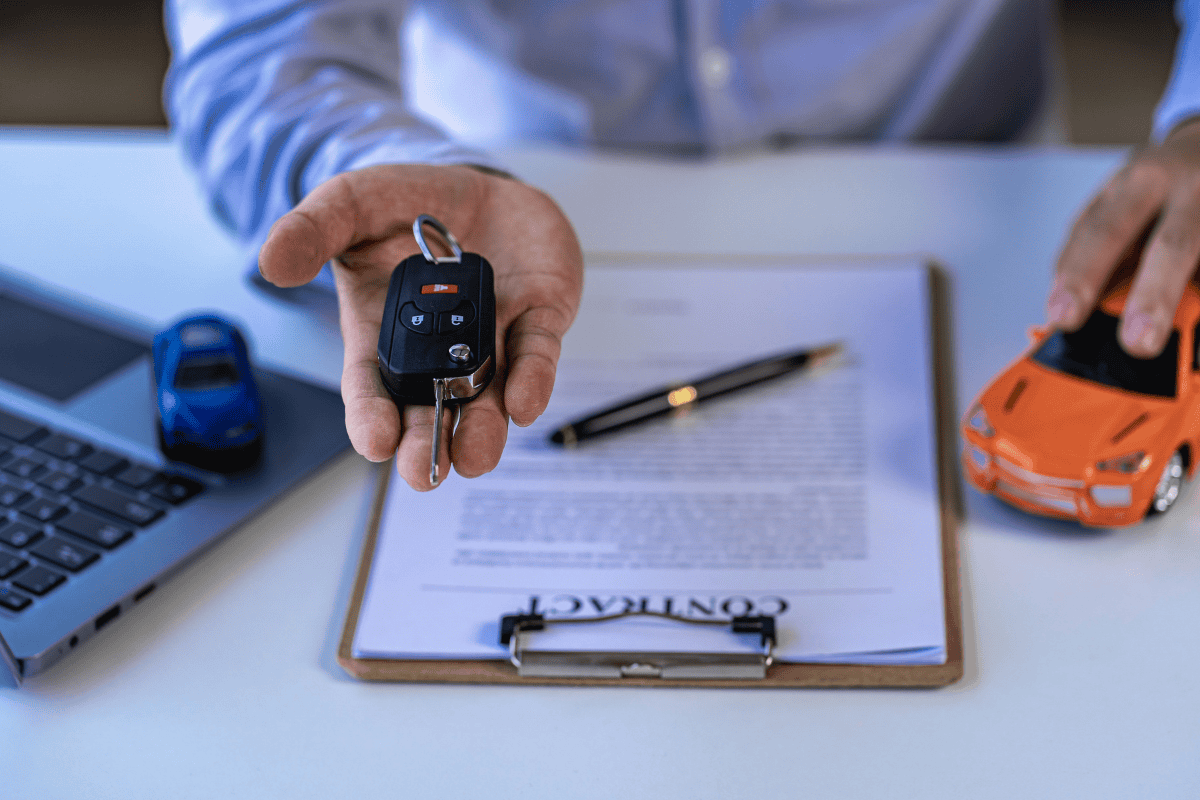 Close-up of a car key fob and keys held over a contract on a clipboard, with a toy car and laptop nearby.