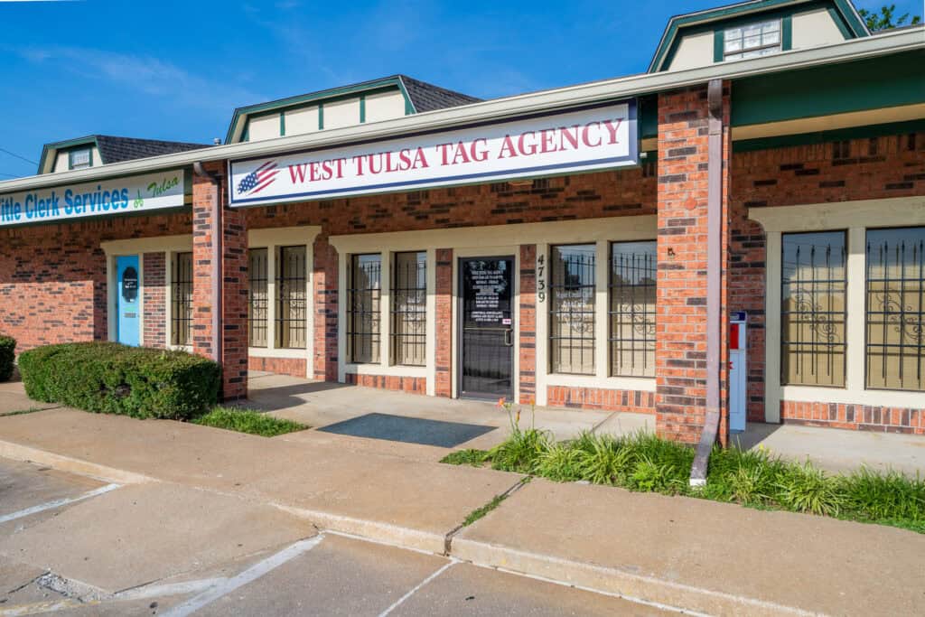 Brick storefront with a 'West Tulsa Tag Agency' sign, green trim, and large windows under a clear blue sky.