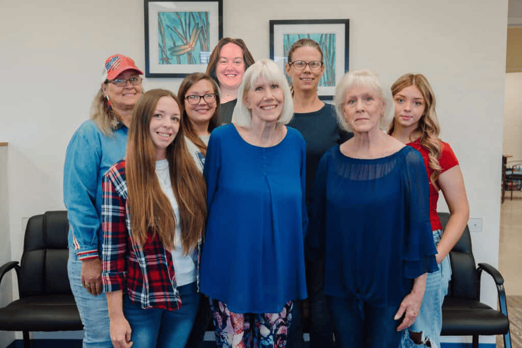 Group of eight women posing together in a bright room, smiling at the camera. Behind them are framed pictures and black chairs.