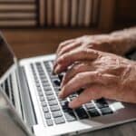 Close-up of wrinkled hands typing on a silver laptop keyboard, with a round-faced wristwatch visible on the left wrist.
