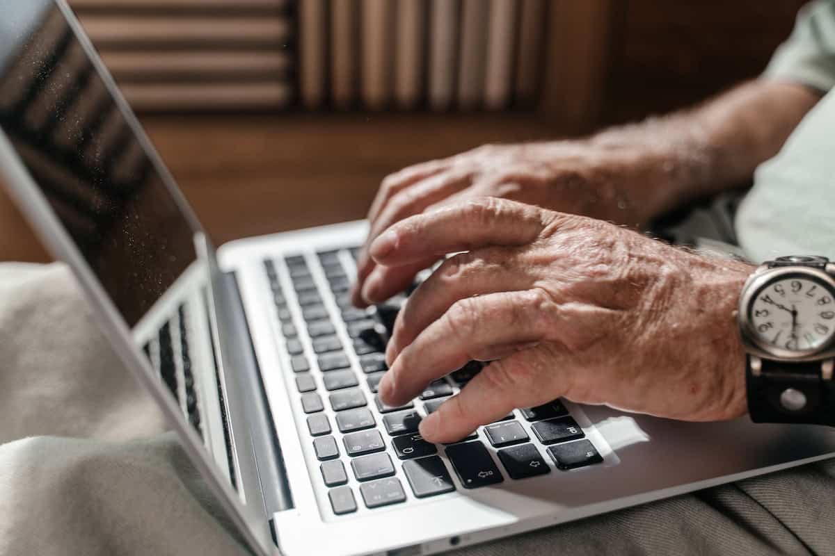 Close-up of wrinkled hands typing on a silver laptop keyboard, with a round-faced wristwatch visible on the left wrist.
